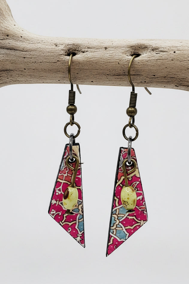A pair of dangling earrings displayed on a clear acrylic stand against a light gray background. The earrings feature geometric triangular charms with a pink, blue, and white patterned background, accented by a small gold circular charm and dark metal fishhook wires.