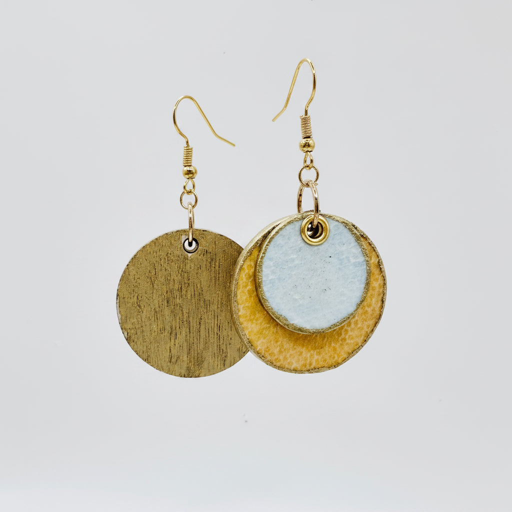 A pair of handmade dangle earrings made from hand painted papers sealed with a matt varnish..  feature a double-tiered circular design with a textured gold-toned outer ring, a light blue-gray textured center disc, and gold-toned fishhook hypoallergenic wires.