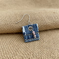 A single handmade dangle earring made from a small square piece of blue denim fabric, with a silver fishhook ear wire, a metal snap closure, and three small pinkish beads, shown next to a black ruler for scale.