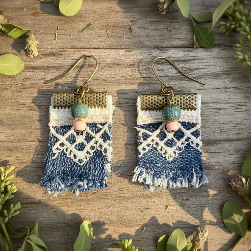 A pair of handmade bohemian-style drop earrings featuring blue denim fringe, vintage white lace applique, textured bronze metal bands, and small blue and pink beads on fishhook wires.