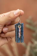 A single dangling earring with a rectangular blue denim-like fabric panel, a silver hook, and a vertical bead strand with pink, white, and silver beads 