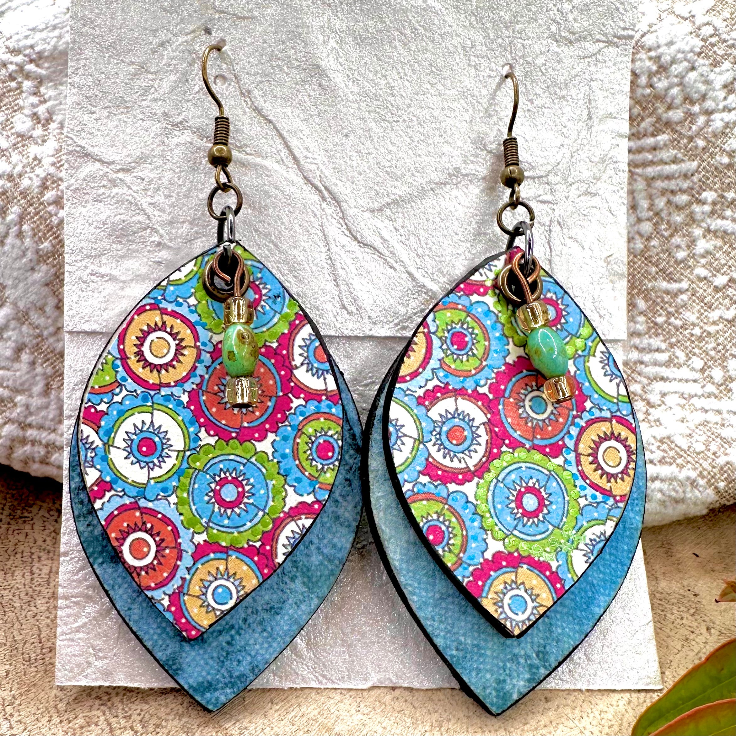 A pair of large teardrop-shaped dangle earrings with a colorful mandala pattern on the front and a textured blue backing, hanging on hypoallergenic vintage brass hooks
