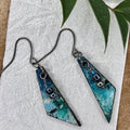 A pair of handmade triangular dangle earrings with a blue and teal marbled pattern, featuring small  bead accents and dark metal hypoallergenic hooks.