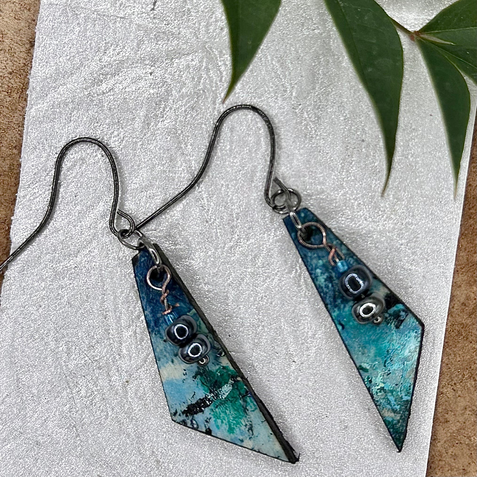 A pair of handmade triangular dangle earrings with a blue and teal marbled pattern, featuring small  bead accents and dark metal hypoallergenic hooks.