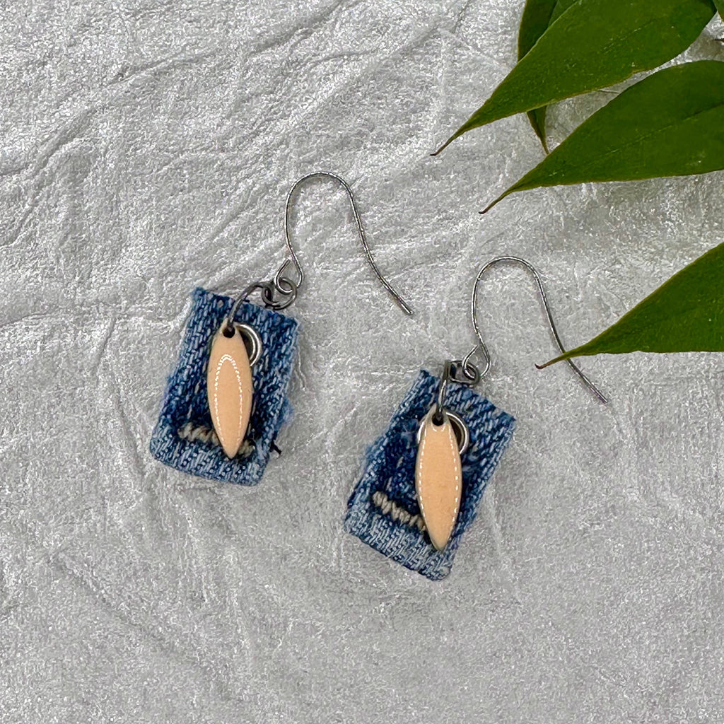 A pair of handmade dangle earrings featuring rectangular blue denim fabric pieces with a smooth, light beige oval shaped charm in the center.  Silver hypoallergenic fishook wires.  .75” without the fishook wire.